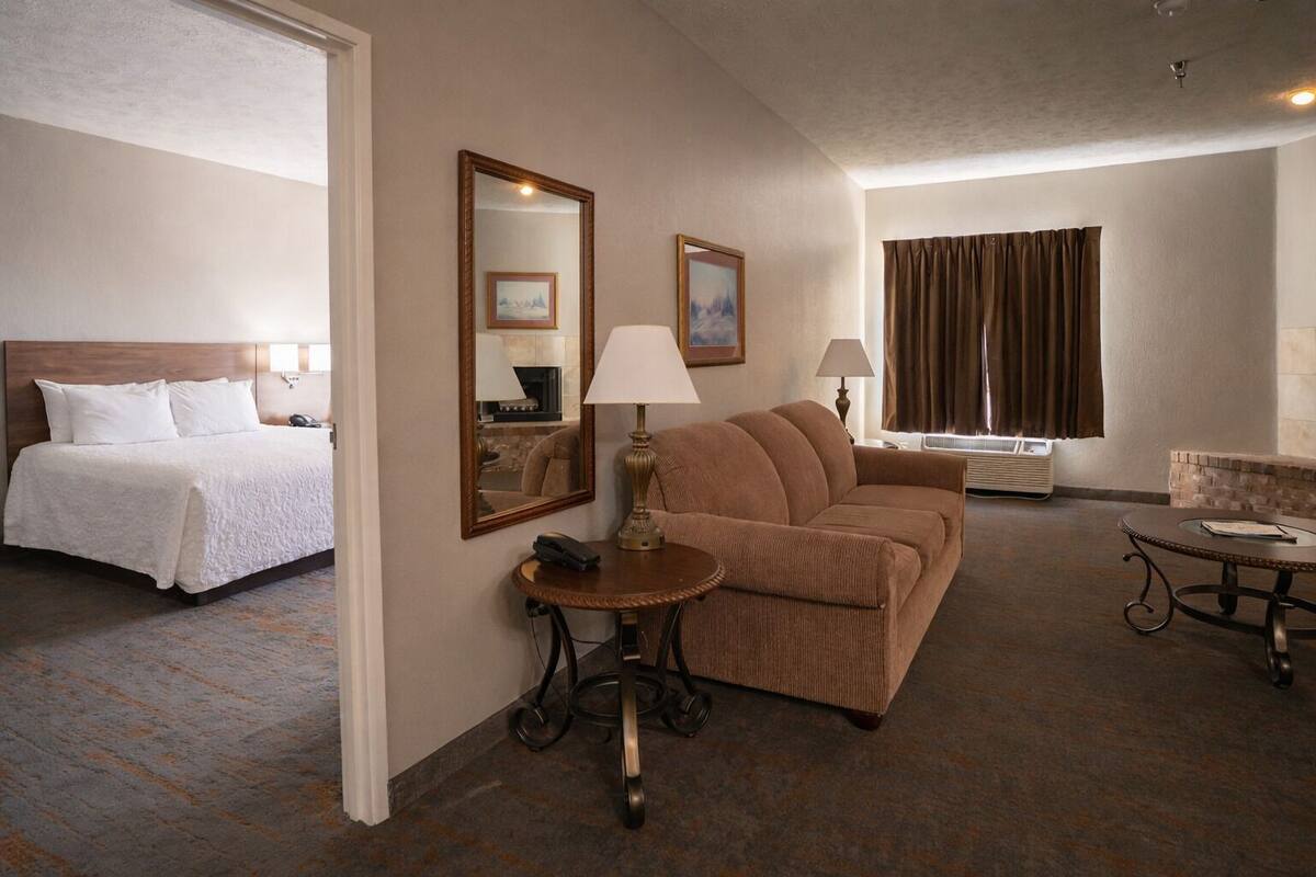 A hotel room with a bed, a sofa, a small table with a lamp, a chair, and a large mirror.
