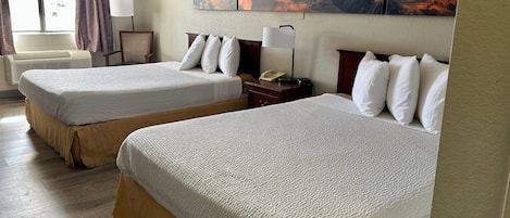 A hotel room with two beds, a large window with curtains, a wall-mounted painting of a canyon, and a bedside table with a lamp.
