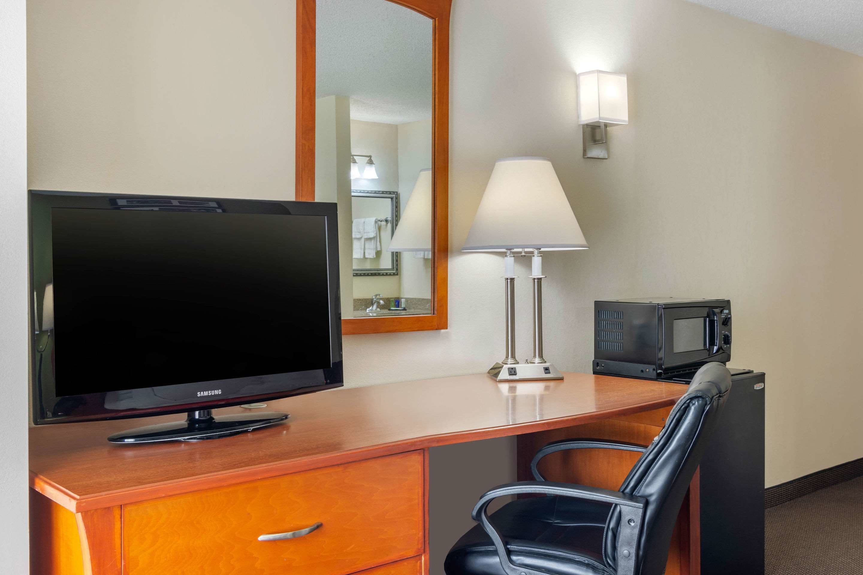 A hotel room with a wooden desk, a black leather chair, a flat-screen TV, a lamp, a microwave, and a mirror.
