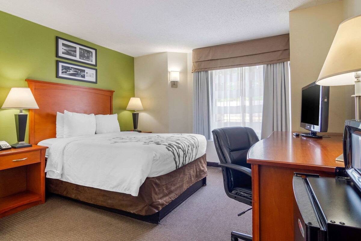 A hotel room with a bed, two bedside tables, a desk with a chair, a television, and a window with curtains.
