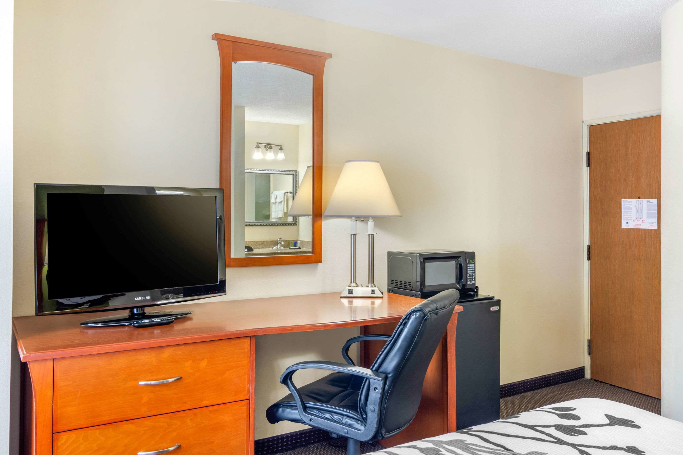 A hotel room with a wooden desk, a television, a mirror, a lamp, a microwave, a chair, and a bed with a patterned bedspread.