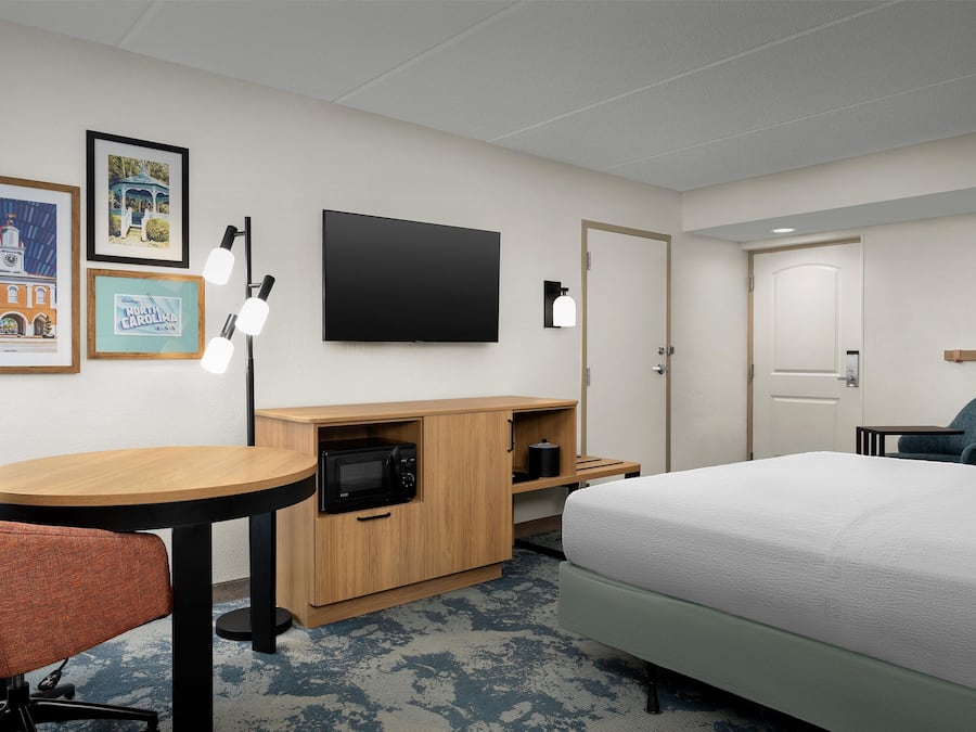 A hotel room with a bed, a desk with a microwave, a chair, a TV, and framed pictures on the wall.