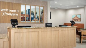A hotel reception area with a wooden counter, a 'Welcome to Hampton' sign, a 'Hilton Honors' plaque, and a view of the cityscape through large windows. - Hampton Inn Fayetteville Fort Bragg (Fayetteville)