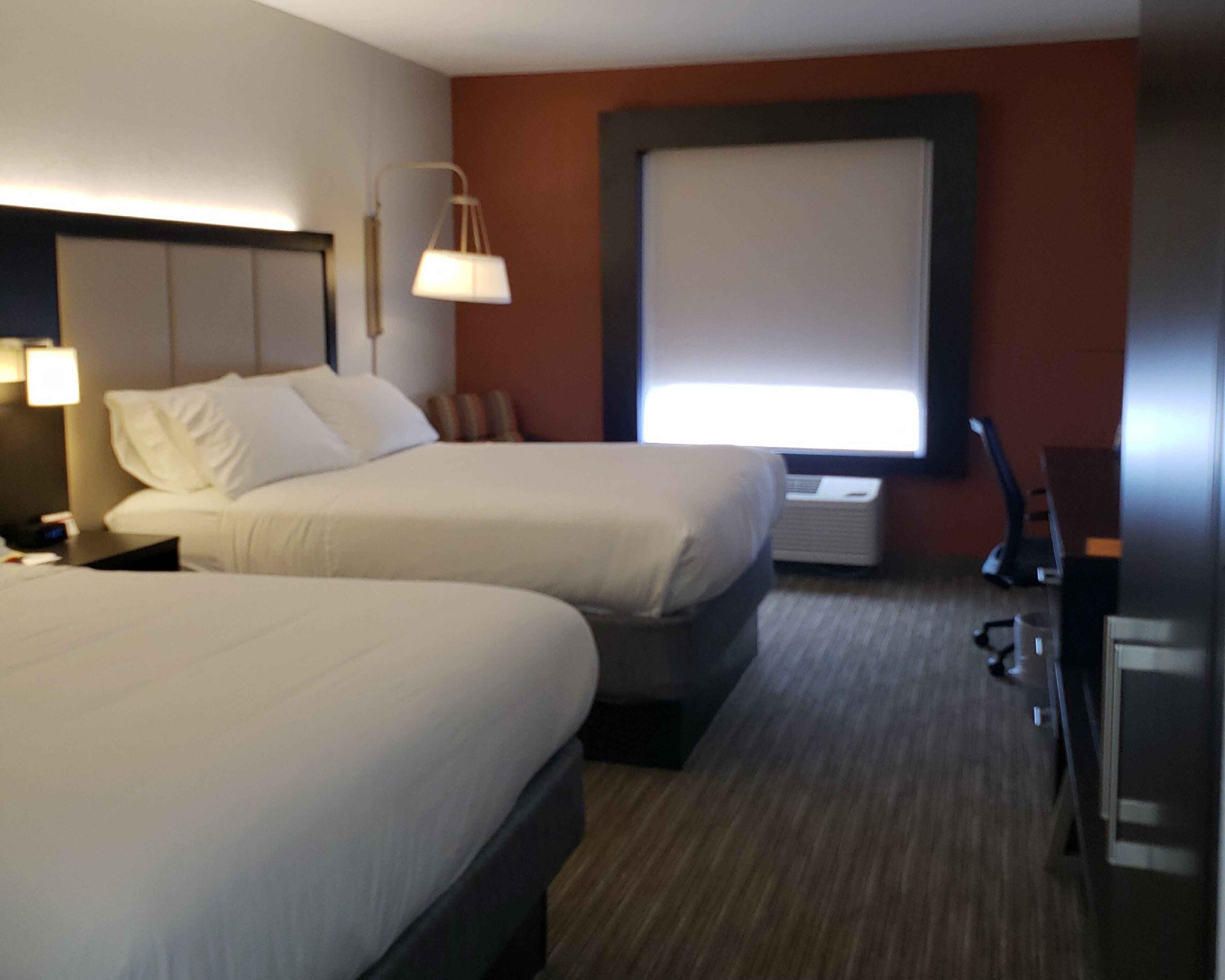 A hotel room with two beds, a desk, a lamp, and a window with blinds.