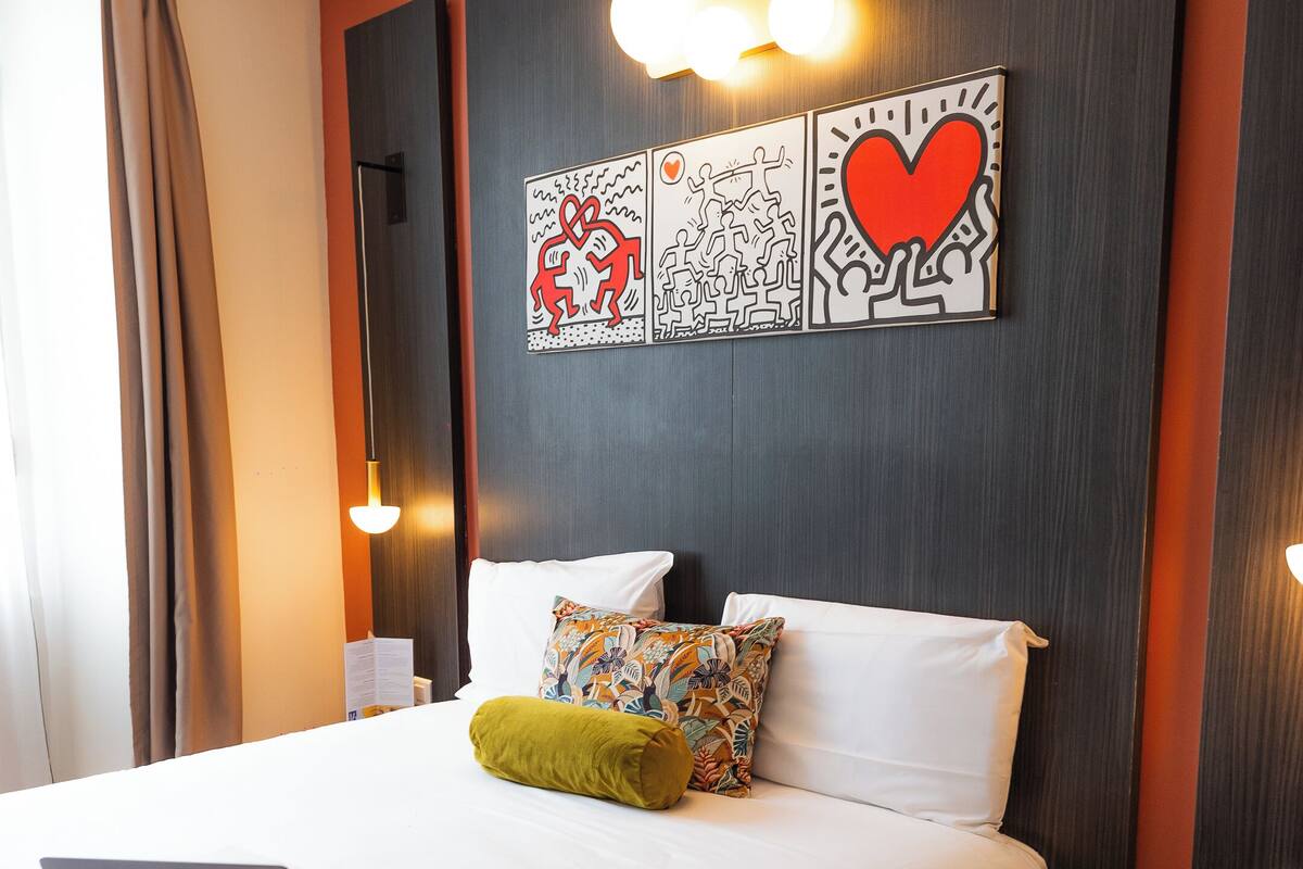 Une chambre d’hôtel avec un lit, un ordinateur portable, un coussin coloré et des œuvres d’art accrochées au mur.