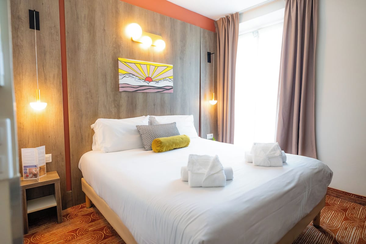 Un lit bien fait, avec du linge de lit blanc et un coussin jaune, une tête de lit en bois agrémentée de lampes fixées au mur, et une fenêtre avec des rideaux.