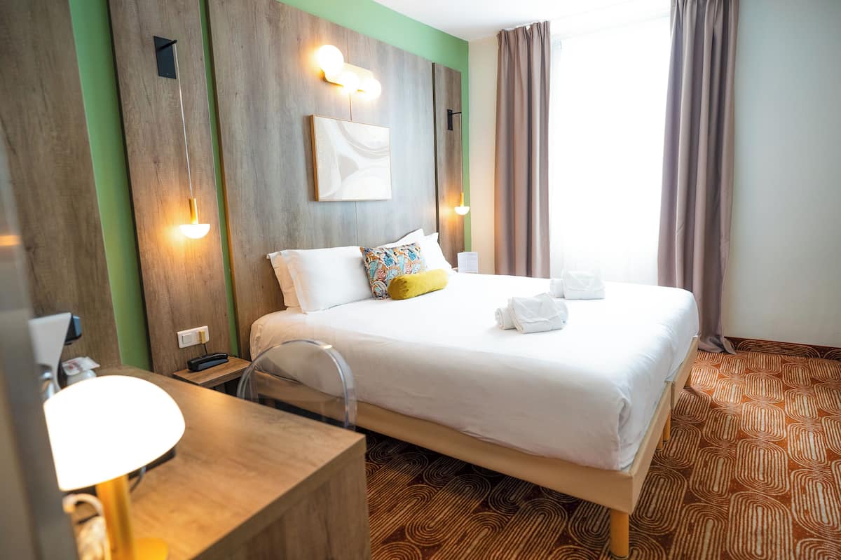 Une chambre d’hôtel avec une tête de lit en bois, un lit avec une literie blanche, une table de chevet avec une lampe et une salle de bain avec un miroir et un lavabo.