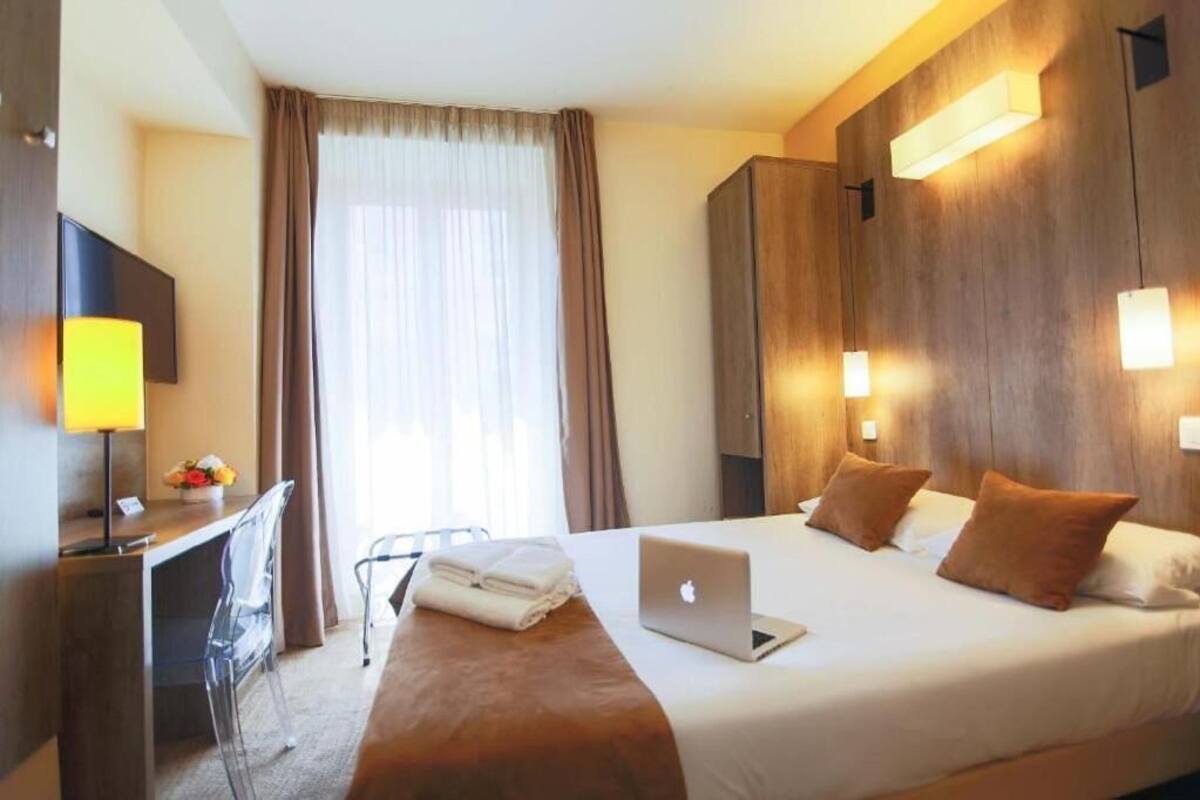 Une chambre d’hôtel avec un lit, un bureau, un ordinateur portable, une chaise et une fenêtre avec des rideaux.