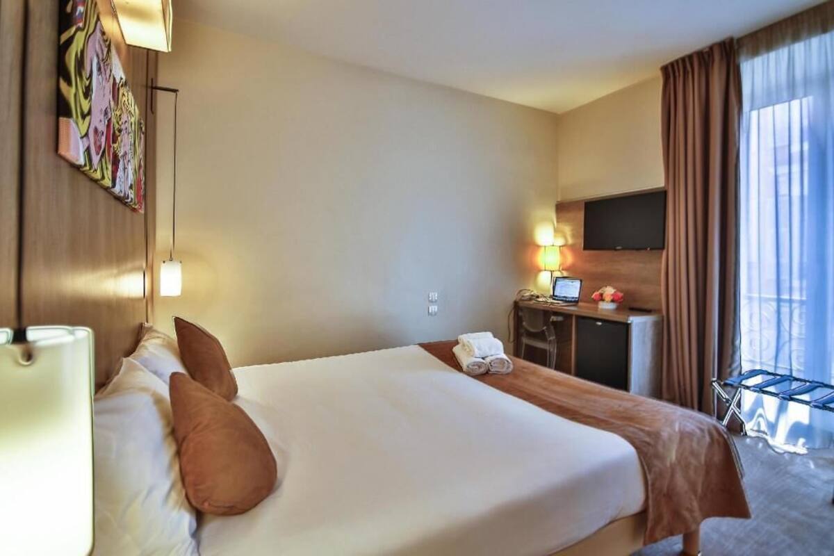 Une chambre d’hôtel avec un lit, un bureau avec un ordinateur, une télévision et une fenêtre avec des rideaux.