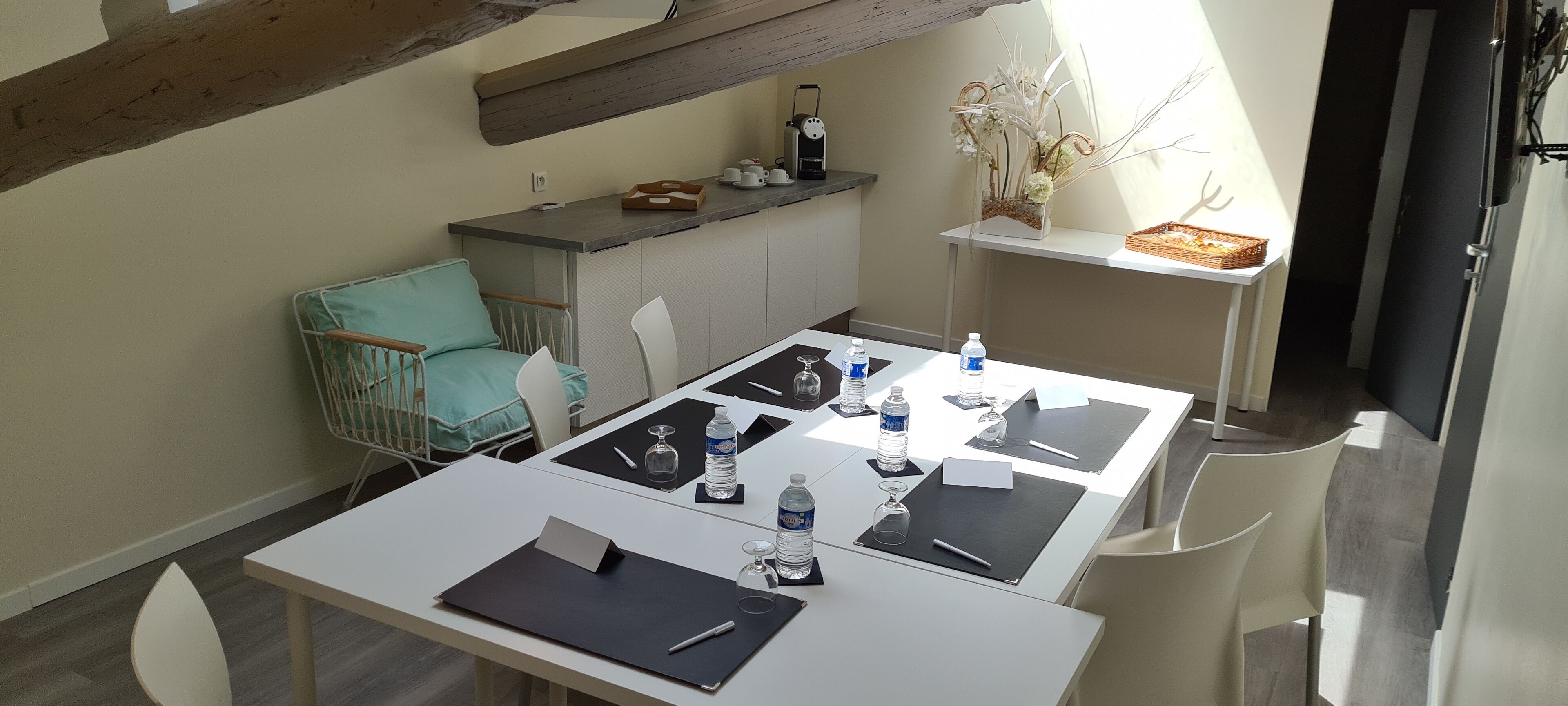 Een eetruimte met een witte tafel gedekt voor vier personen, voorzien van waterflessen, glazen en een notitieboekje. Een blauwgroene stoel staat bij de tafel, en een klein kastje met keukengerei is op de achtergrond zichtbaar.