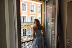 Una mujer parada en un balcón, observando una hilera de edificios con balcones y contraventanas.