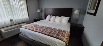 A hotel room with a large bed, a bedside table with a lamp, a heater, and a window with blinds.