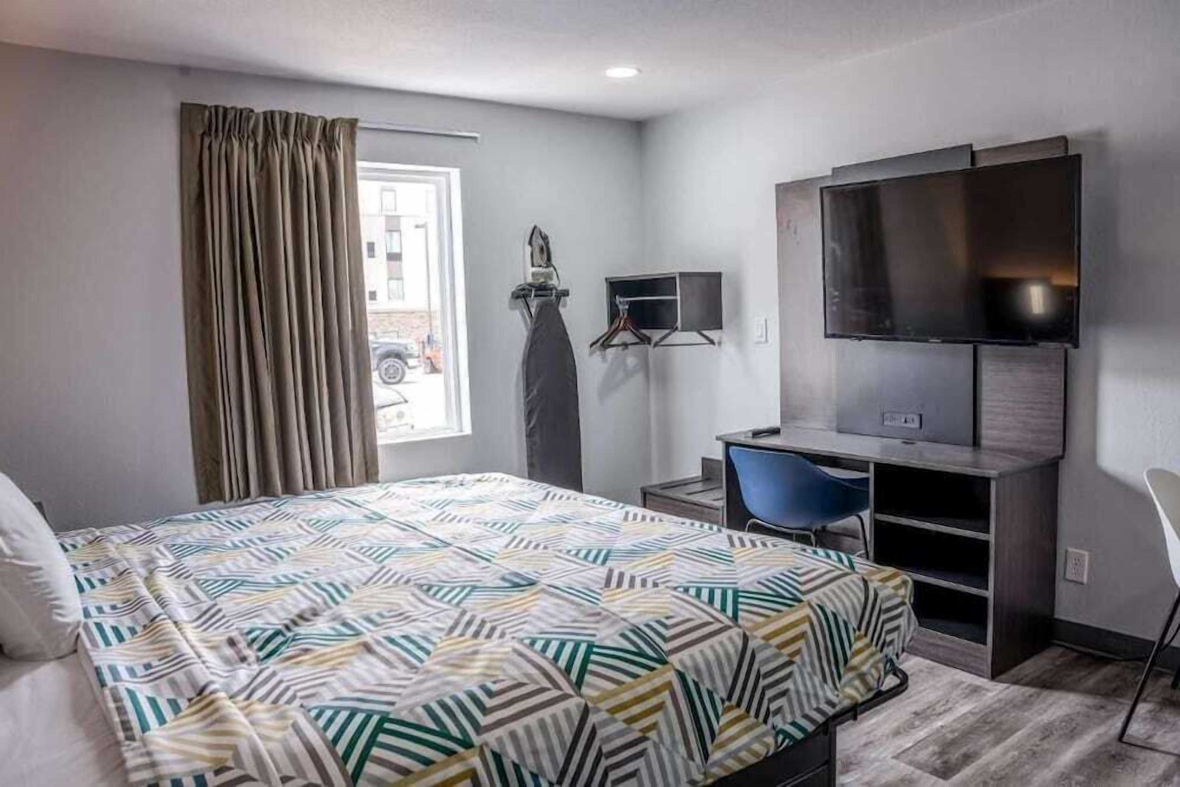 Une chambre d’hôtel comprenant un lit, un bureau avec une chaise, une télévision et une fenêtre avec des rideaux.