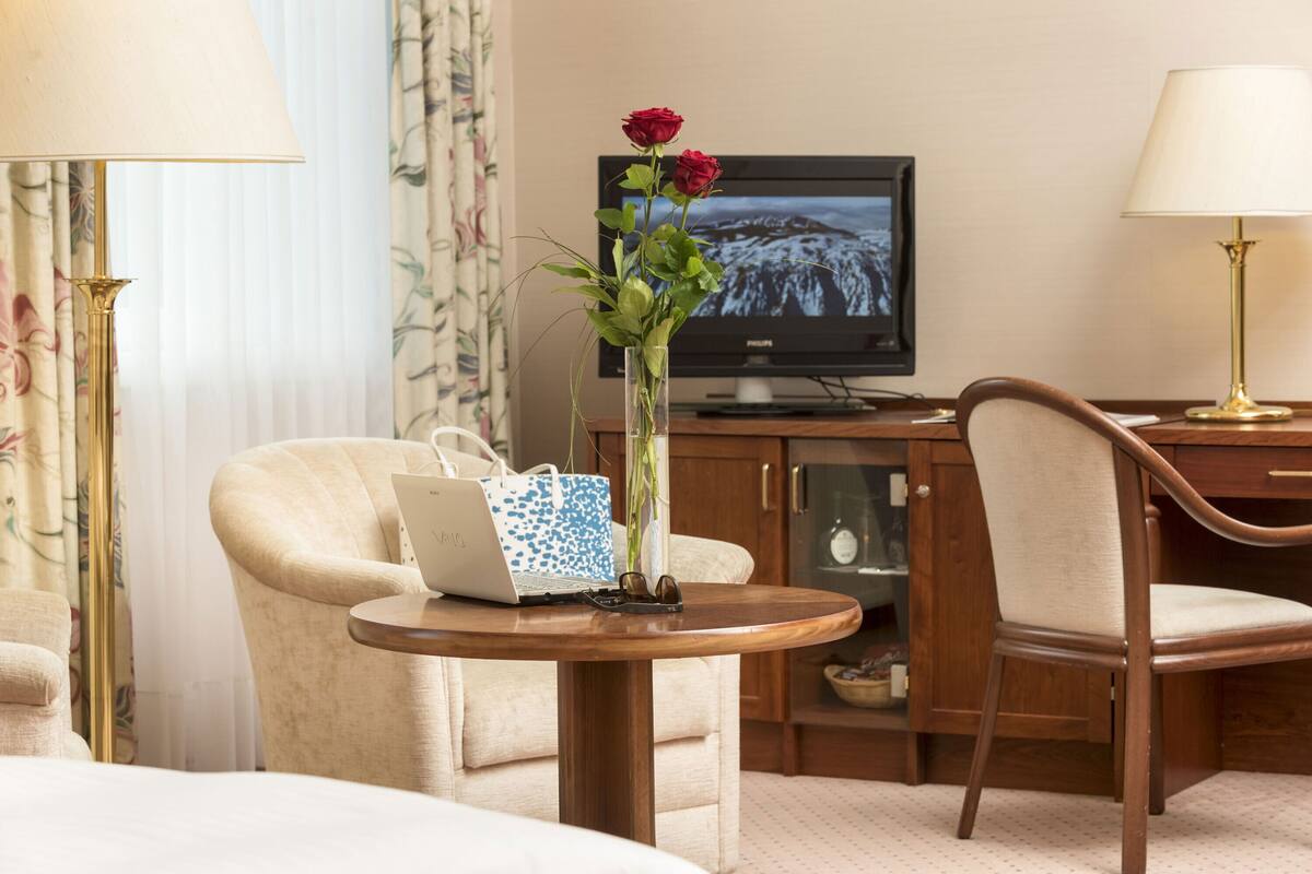 A hotel room with a bed, a desk with a laptop, a vase with flowers, a television, and a chair.