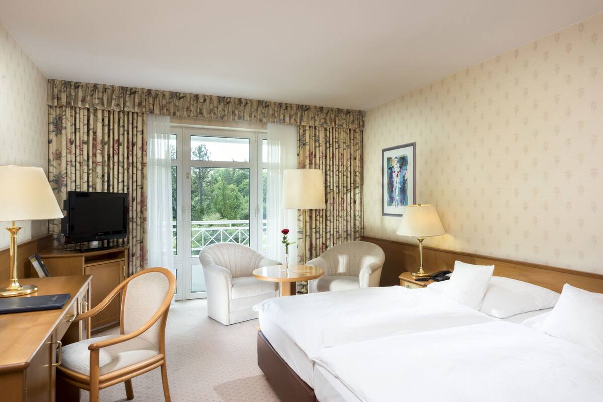 A hotel room with a bed, two chairs, a desk with a lamp, a television, and a window with curtains.