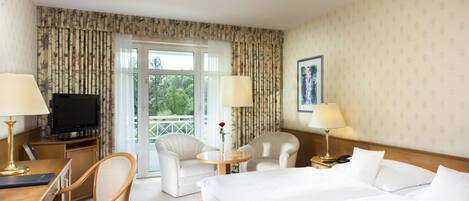 Una habitación de hotel con una cama, dos sillas, un escritorio con lámpara, un televisor y una ventana con cortinas.