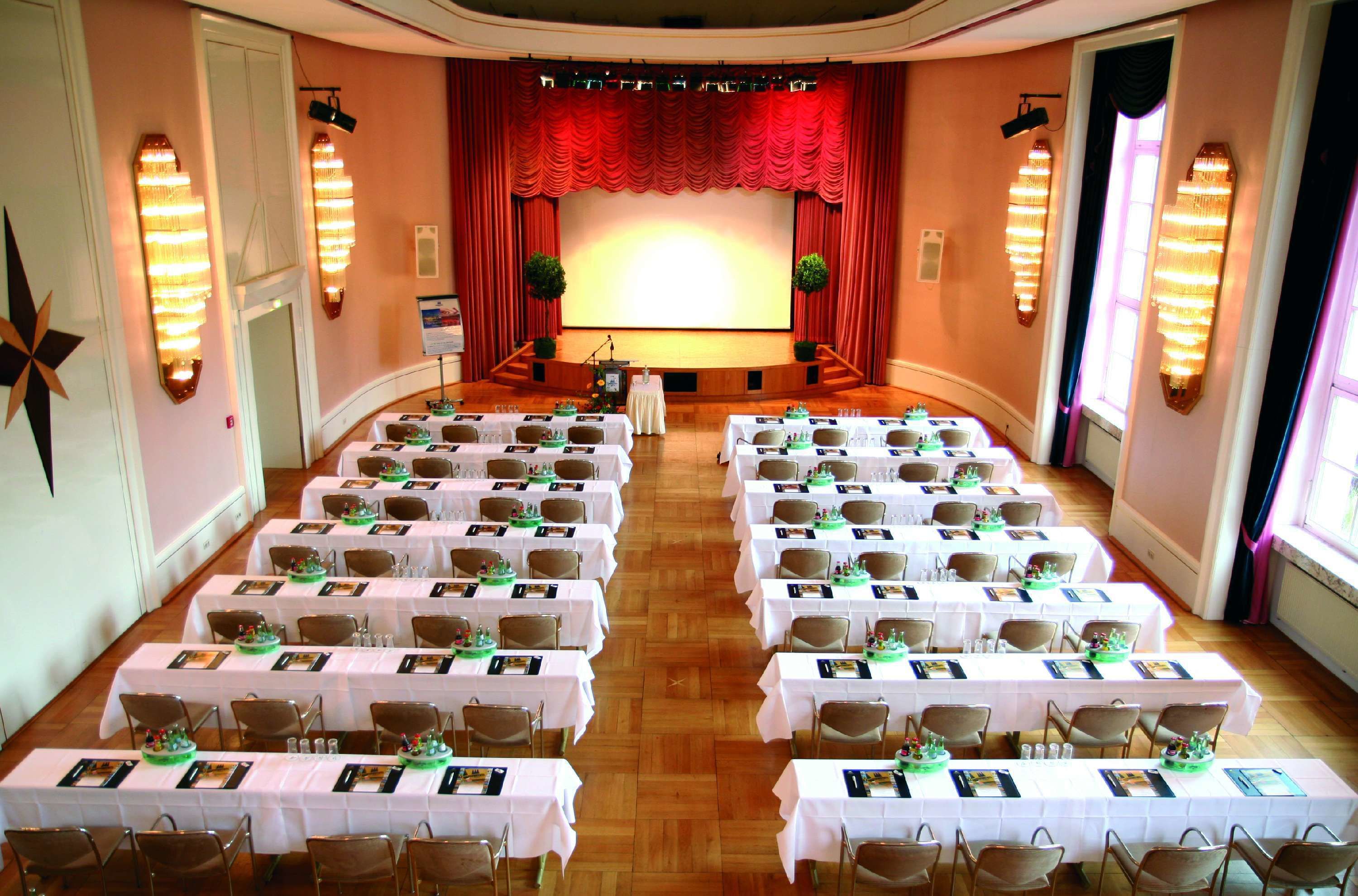 A conference room with rows of tables set for a meeting, a stage with curtains, and wall-mounted lights.