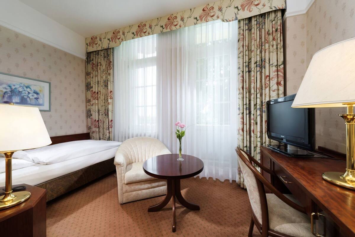 A hotel room with a bed, a chair, a round table with a vase, a desk with a lamp, and a television.