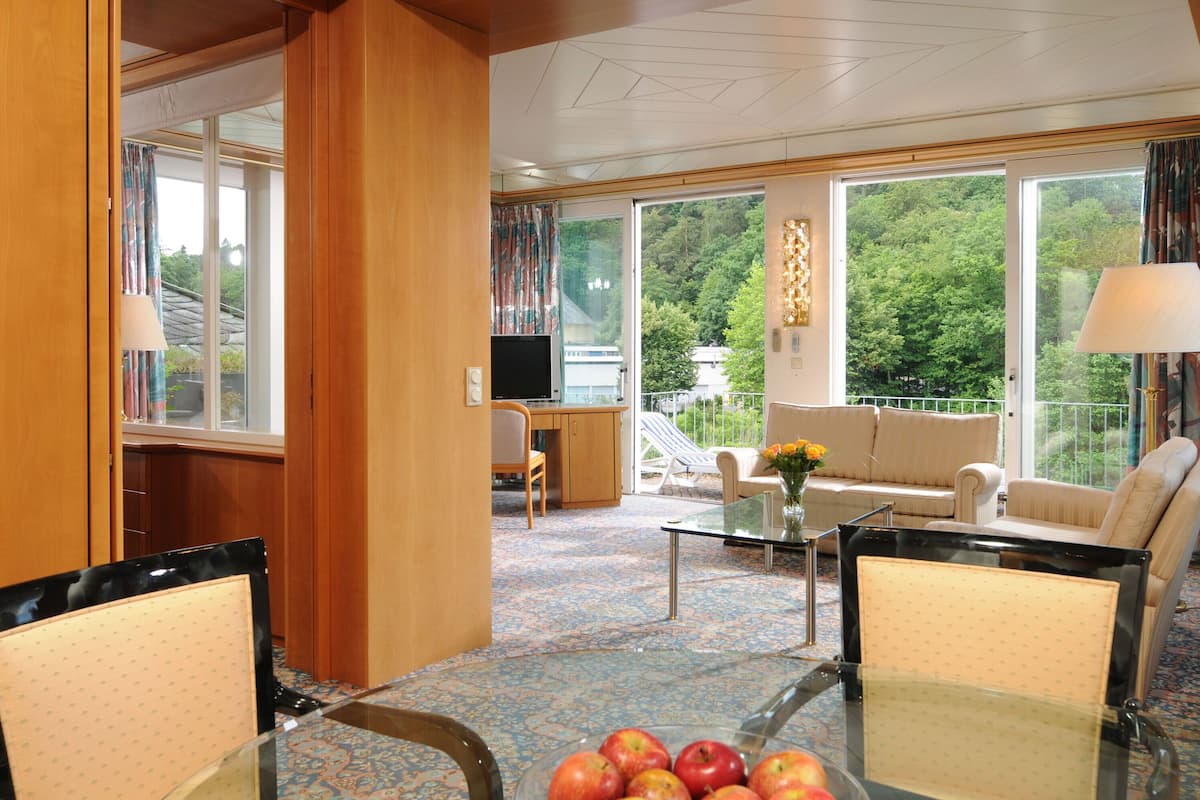 A hotel room with a sofa, chairs, a TV, a glass table with apples, and large windows with a view of trees.