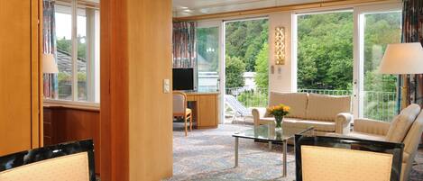 A hotel room with a sofa, chairs, a TV, a glass table with apples, and large windows with a view of trees.