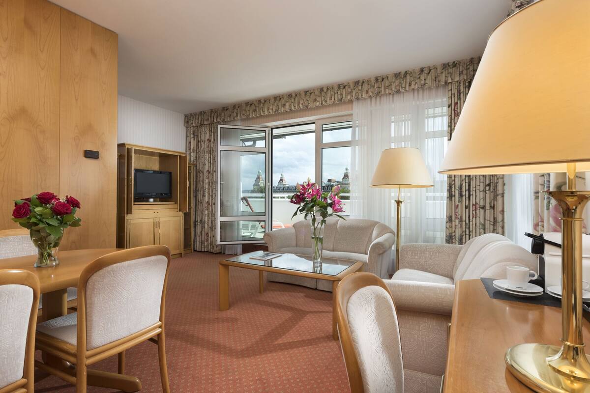 A hotel room with a wooden wardrobe, a television, a sofa, a coffee table with flowers, and a view of a cityscape through large windows.