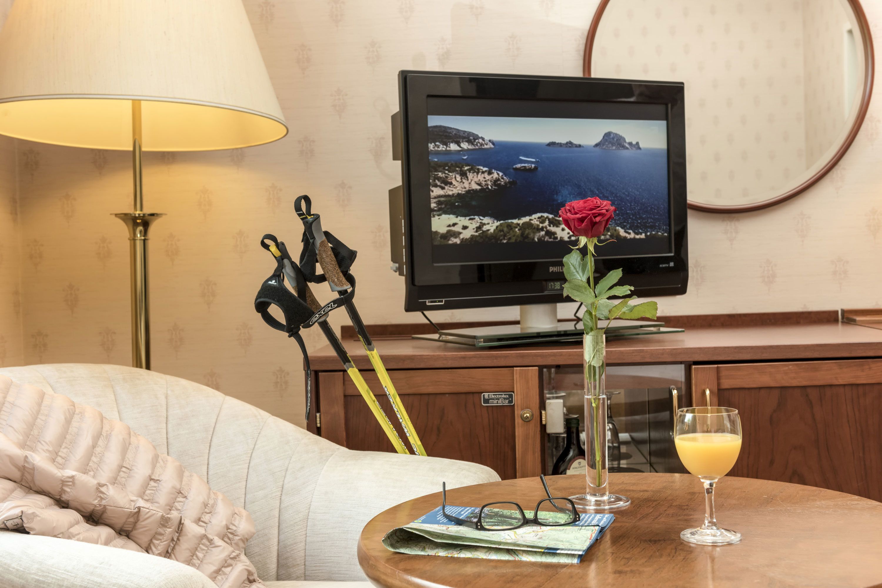 Quarto de hotel com televisão exibindo uma vista panorâmica, uma mesa com uma rosa, copos e uma bebida, e um abajur.
