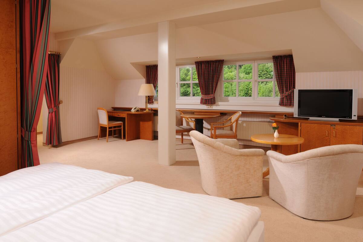 A hotel room with a bed, two armchairs, a desk with a lamp, a television, and a window with curtains.