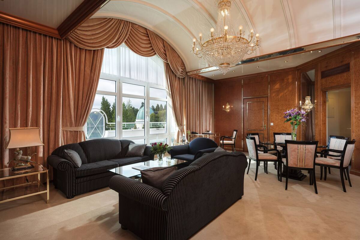 A spacious living room with a large window, a chandelier, and a dining area with chairs and a table.
