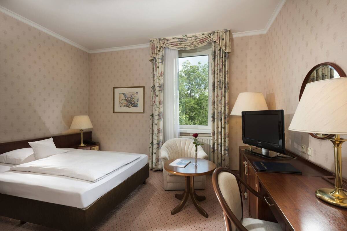 A hotel room with a bed, a desk with a TV, a chair, a small table with a book and a vase with a flower, and a window with curtains.