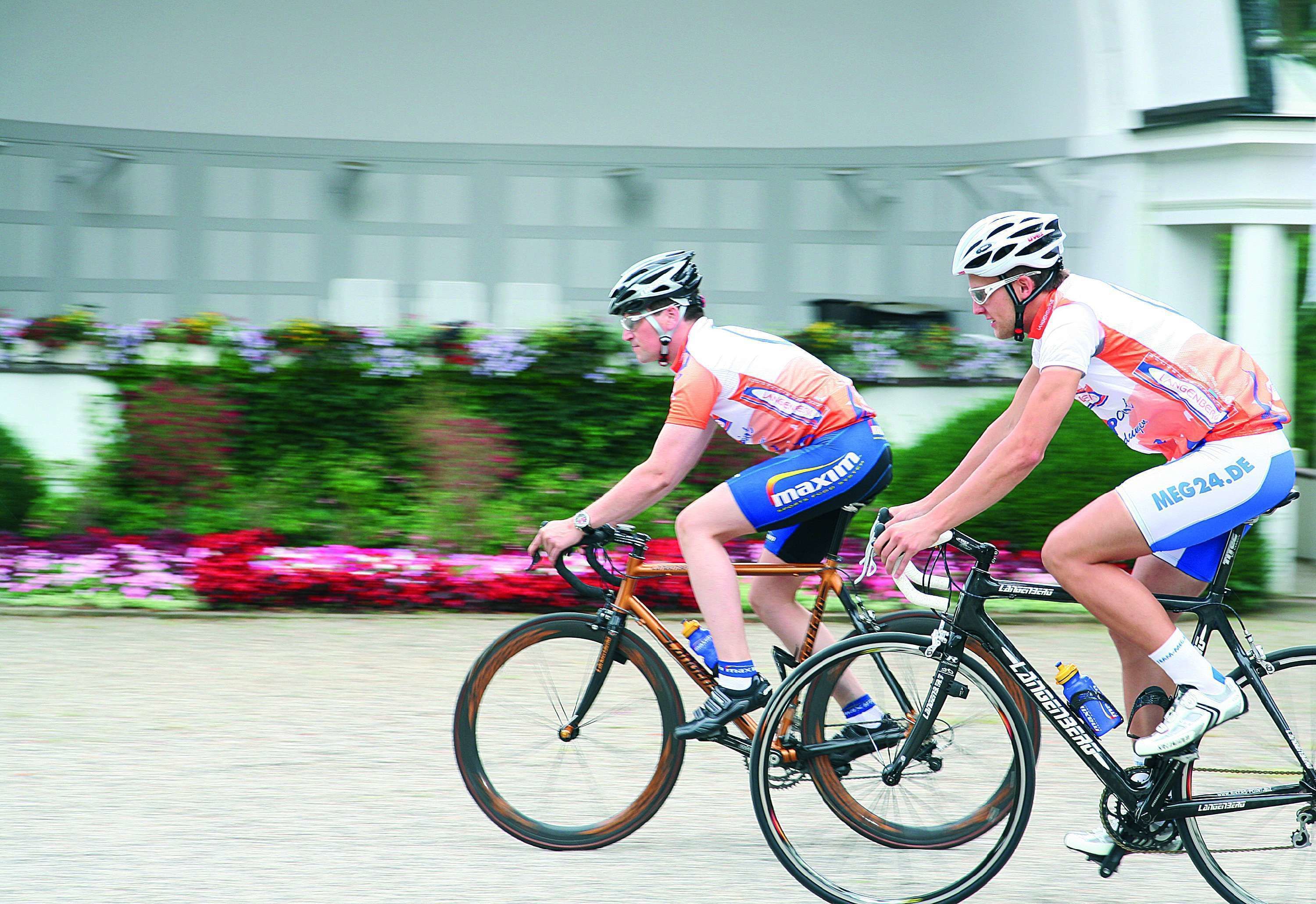 Dos ciclistas en una carretera con un edificio y macizos de flores al fondo.