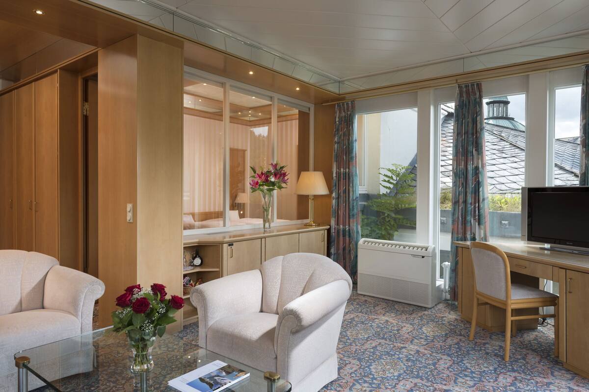A hotel room with a sofa, armchair, desk, TV, and a large window with curtains.