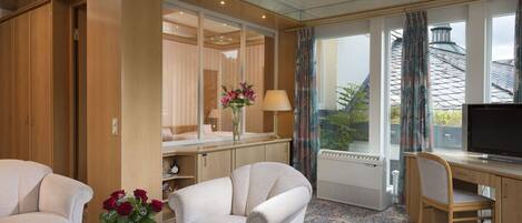 Habitación de hotel con sofá, sillón, escritorio, televisor y un amplio ventanal con cortinas.