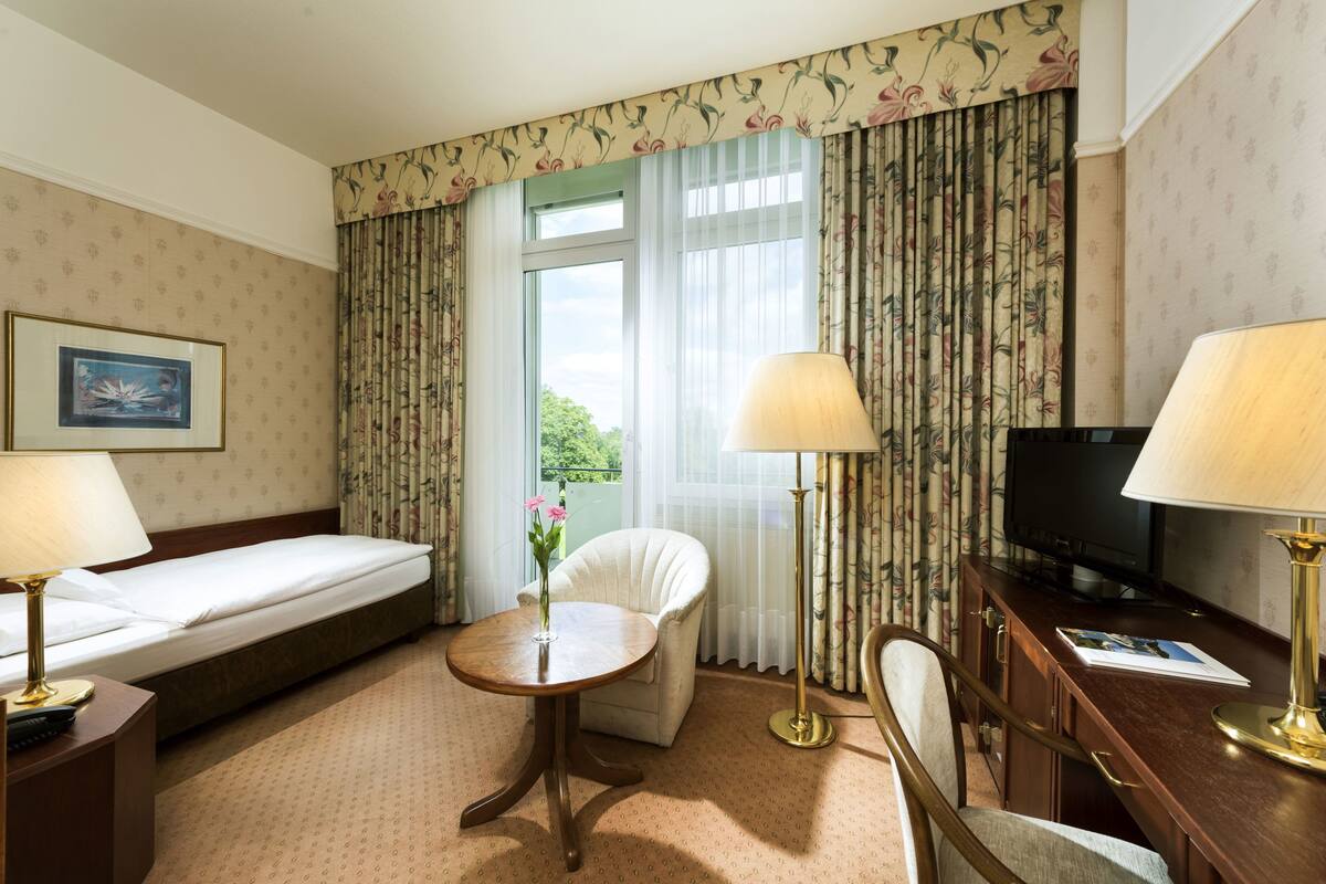A hotel room with a bed, a desk with a television, a chair, a table with a vase of flowers, and curtains with a floral pattern.