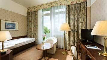 Una habitación de hotel con cama, escritorio con televisor, silla, pequeña mesa con un florero y cortinas.