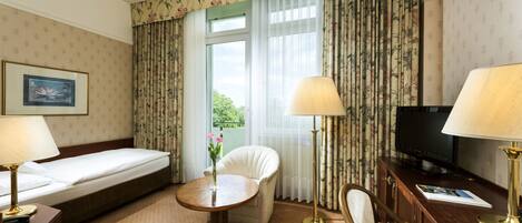 Una habitación de hotel con cama, escritorio con televisor, silla, pequeña mesa con un florero y cortinas.