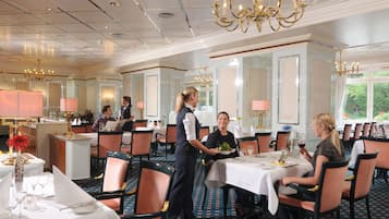 A formal dining area with tables set for multiple guests, a waiter attending to a table, and a view of the outdoors through large windows.