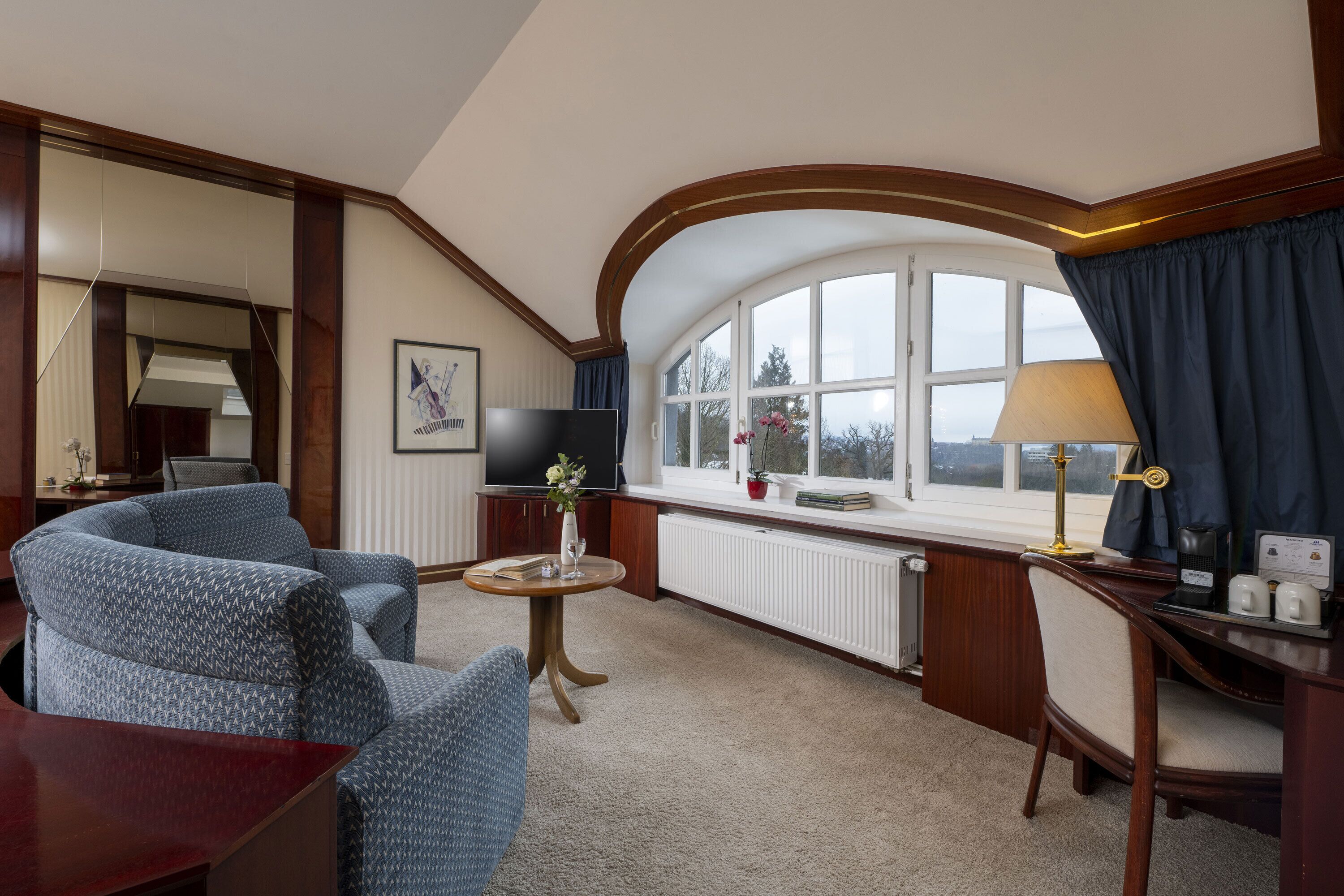 Ein Hotelzimmer mit gewölbter Decke, einem großen Fenster mit Vorhängen, einem Flachbildfernseher, einem kleinen runden Tisch mit einer Vase sowie einem Schreibtisch mit einer Lampe.