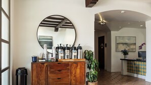 Un meuble en bois avec un miroir, une poubelle et une plante en pot, situé dans un couloir dont le sol est recouvert d’un motif décoratif et où l’on aperçoit une porte au fond.