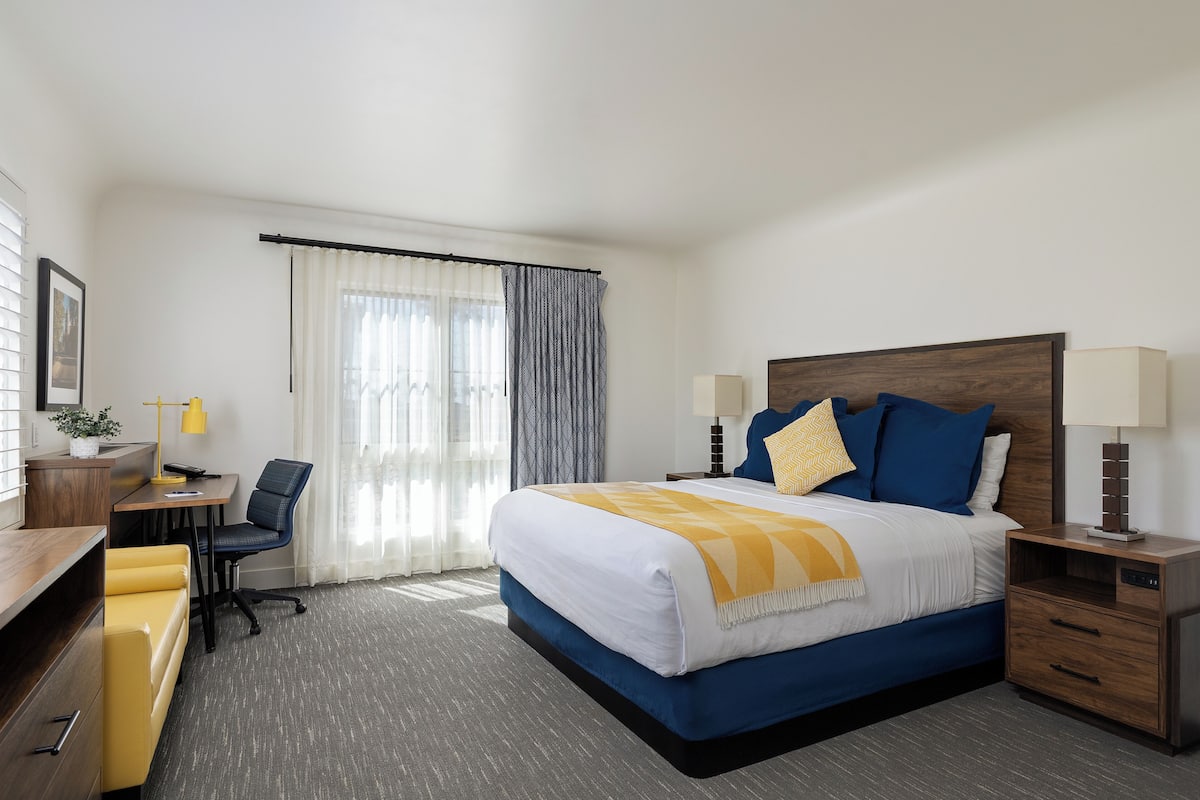 Quarto de hotel com cama, escrivaninha, cadeira e mesa de cabeceira com luminárias.