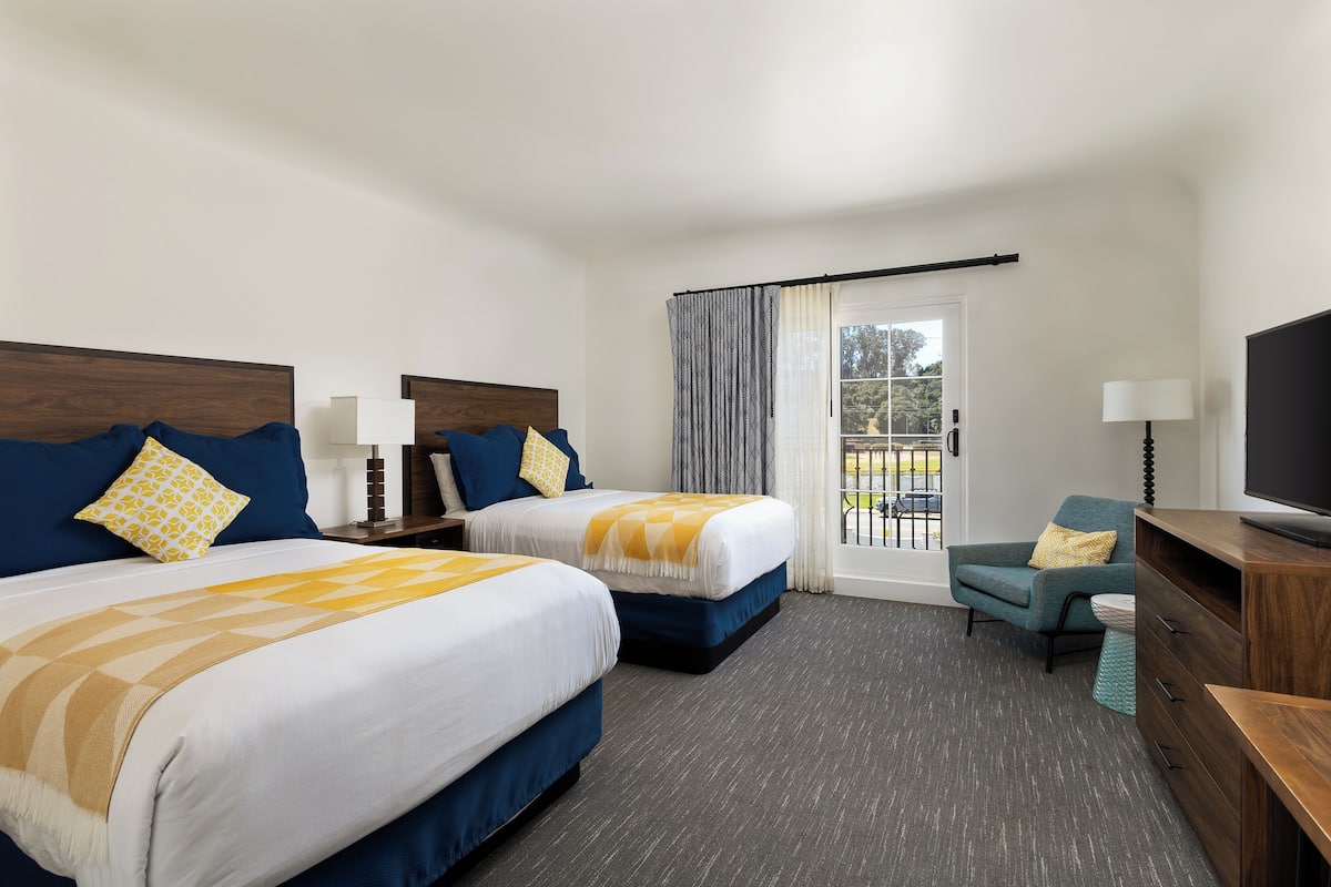 Quarto de hotel com duas camas, televisão, cadeira e varanda com cortinas.