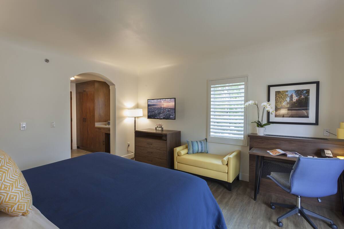 Quarto de hotel com cama, escrivaninha, cadeira, sofá, televisão e janela com persianas.