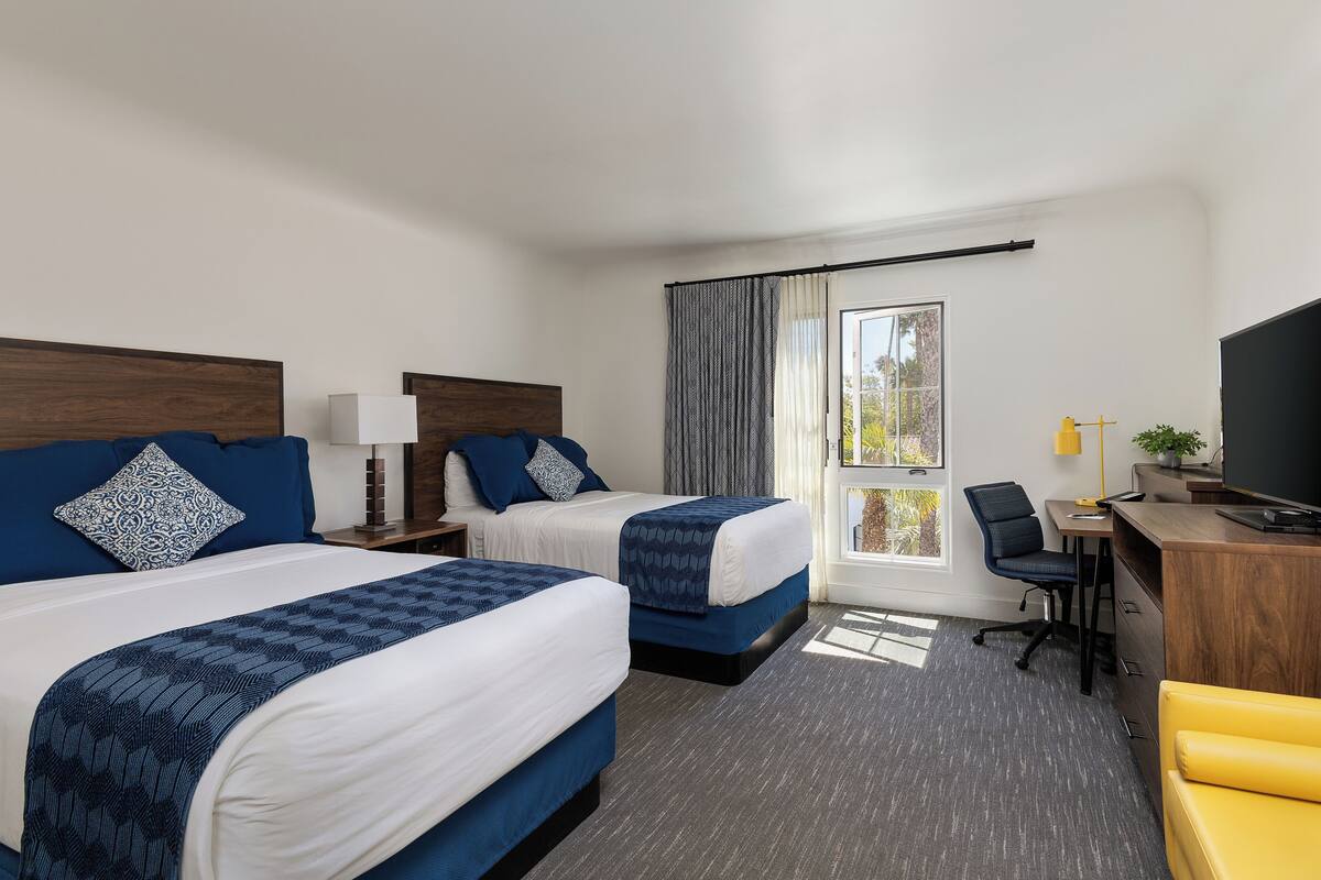 Quarto de hotel com duas camas, uma escrivaninha, uma cadeira, uma televisão e uma janela com cortinas.