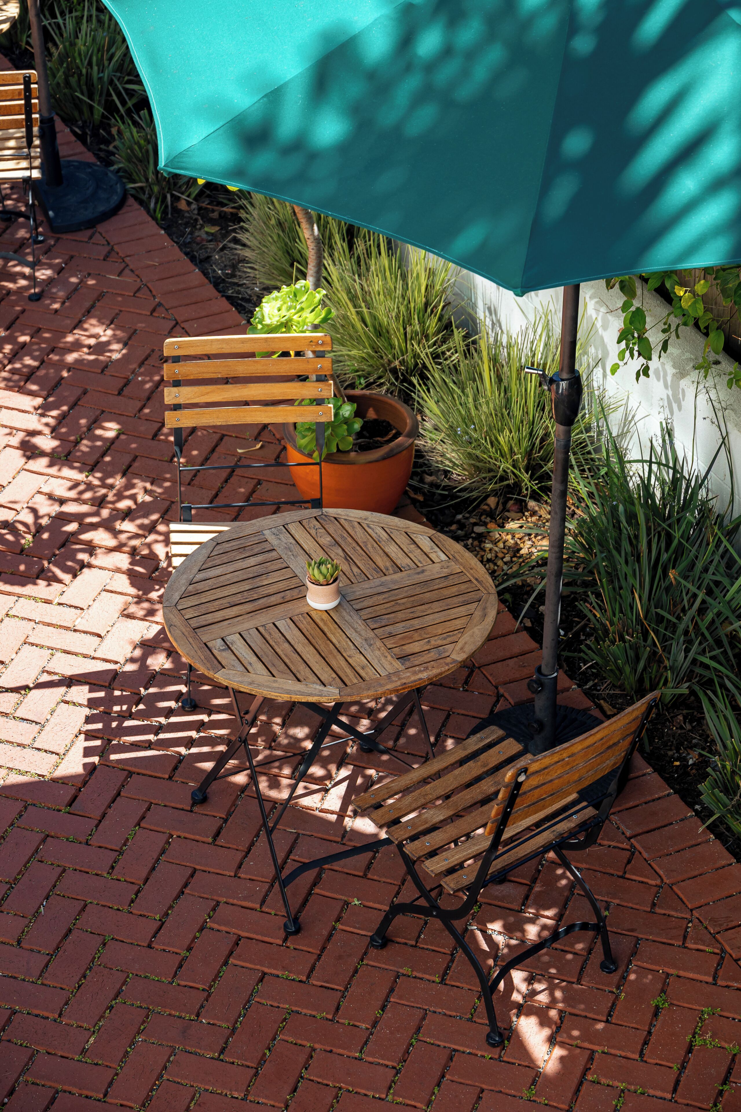 A patio with a wooden table and chairs, a potted plant, and a green umbrella.