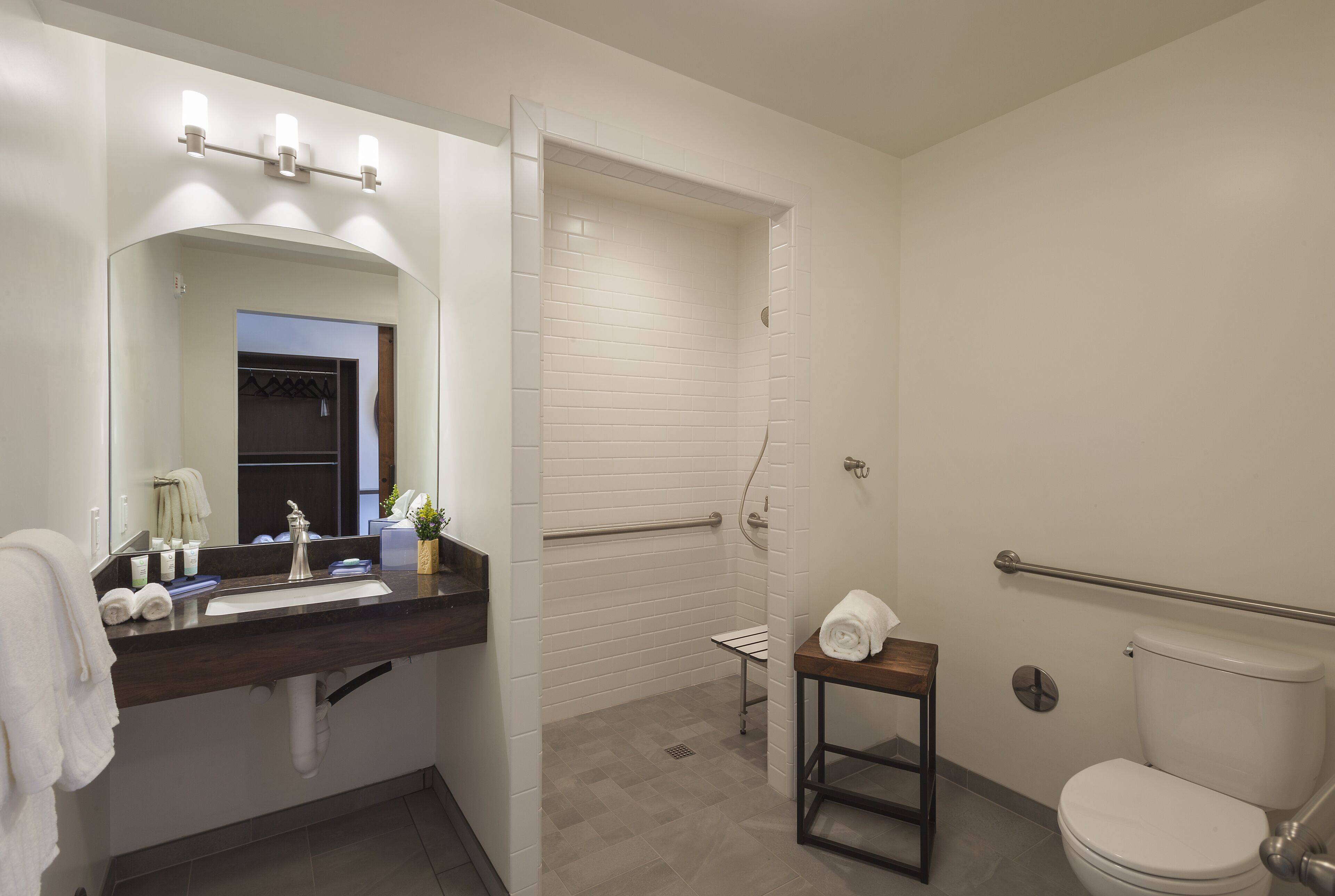 Un baño moderno con encimera oscura, baldosas blancas, un espejo grande, un inodoro y una zona de ducha.