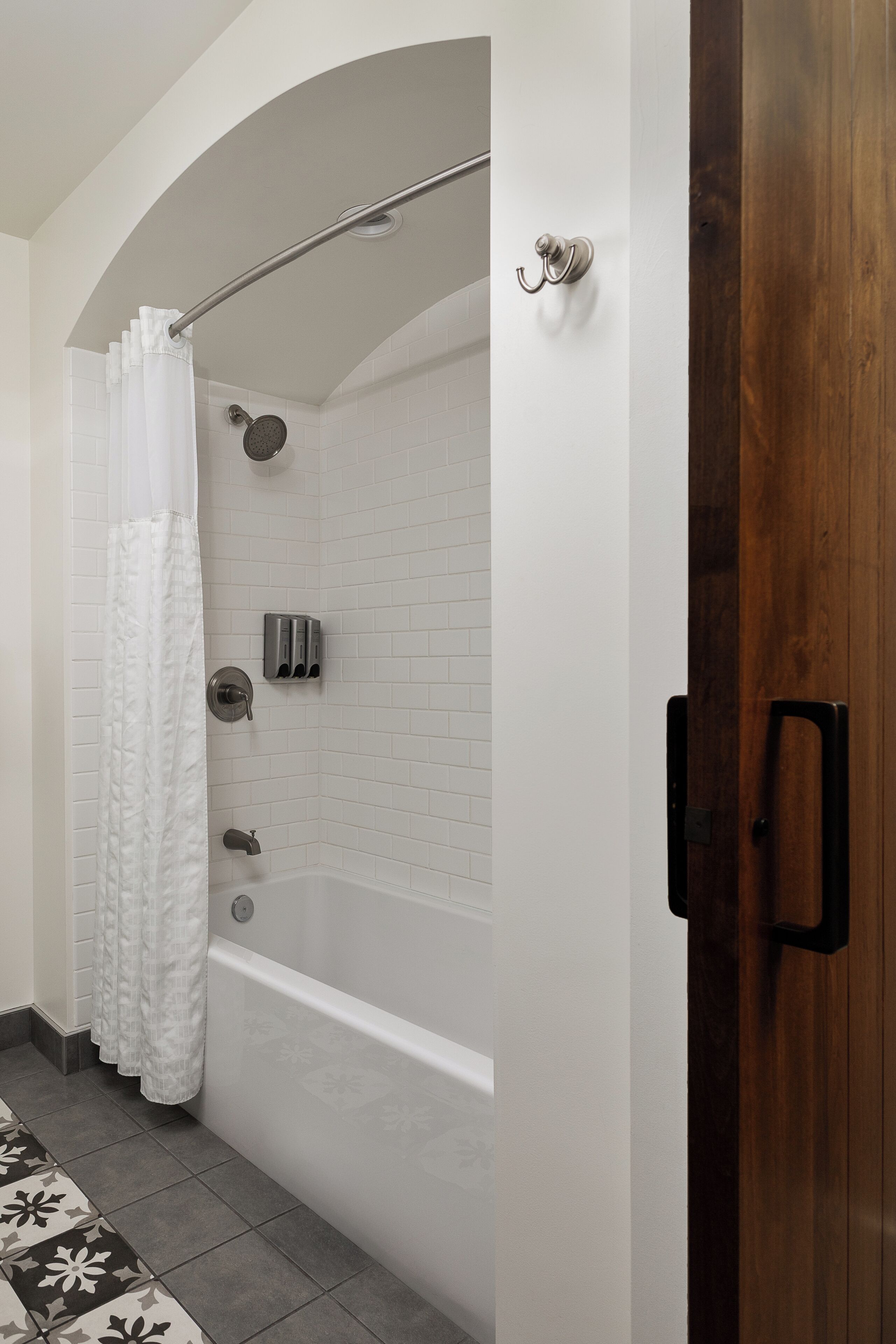 Een badkamer met een douche, een bad, witte betegelde wanden, een houten deur en een vloer met een patroon.