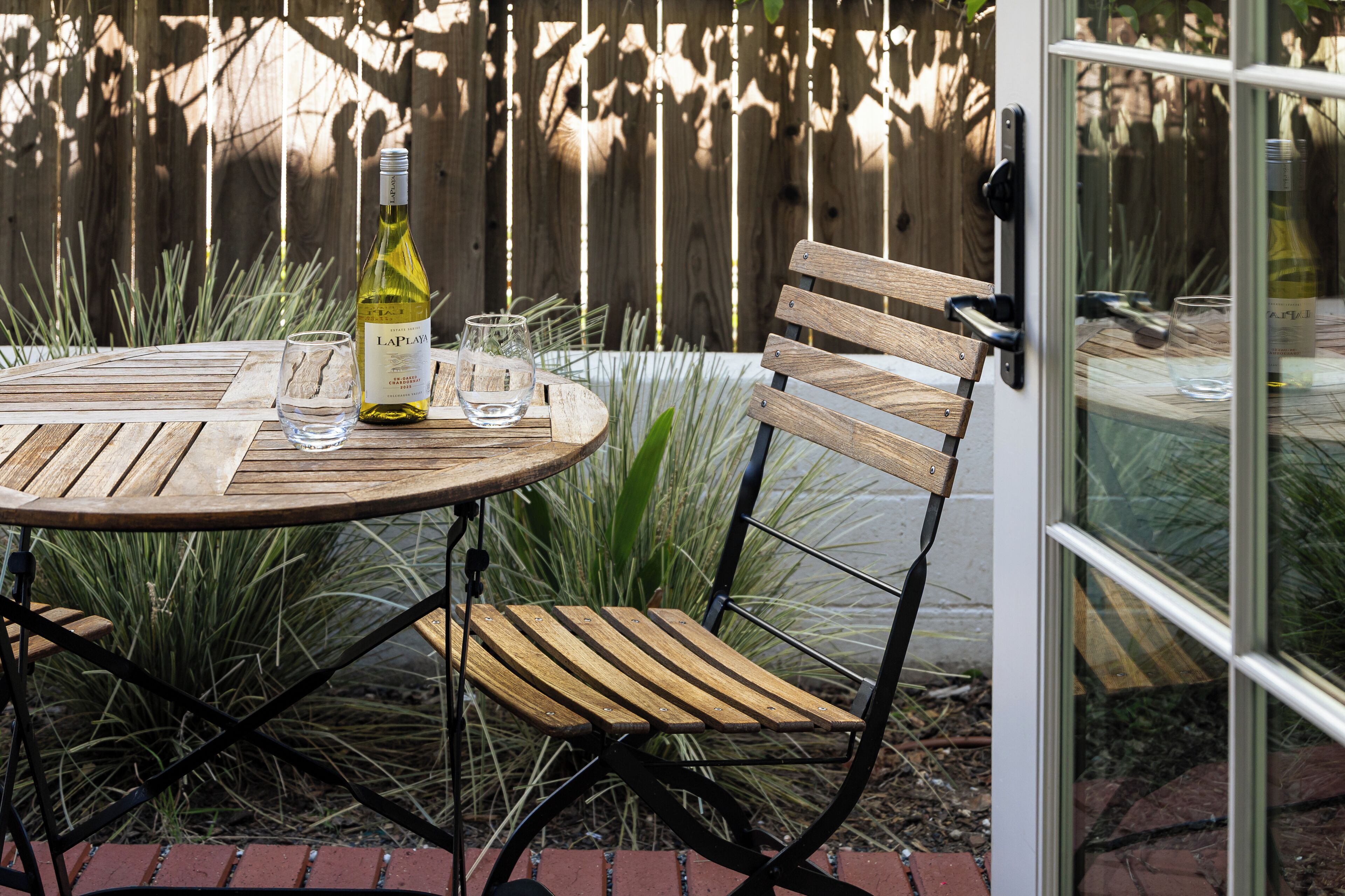 A round wooden table with a bottle of wine and glasses, a wooden chair, and a glass door with a view of greenery.