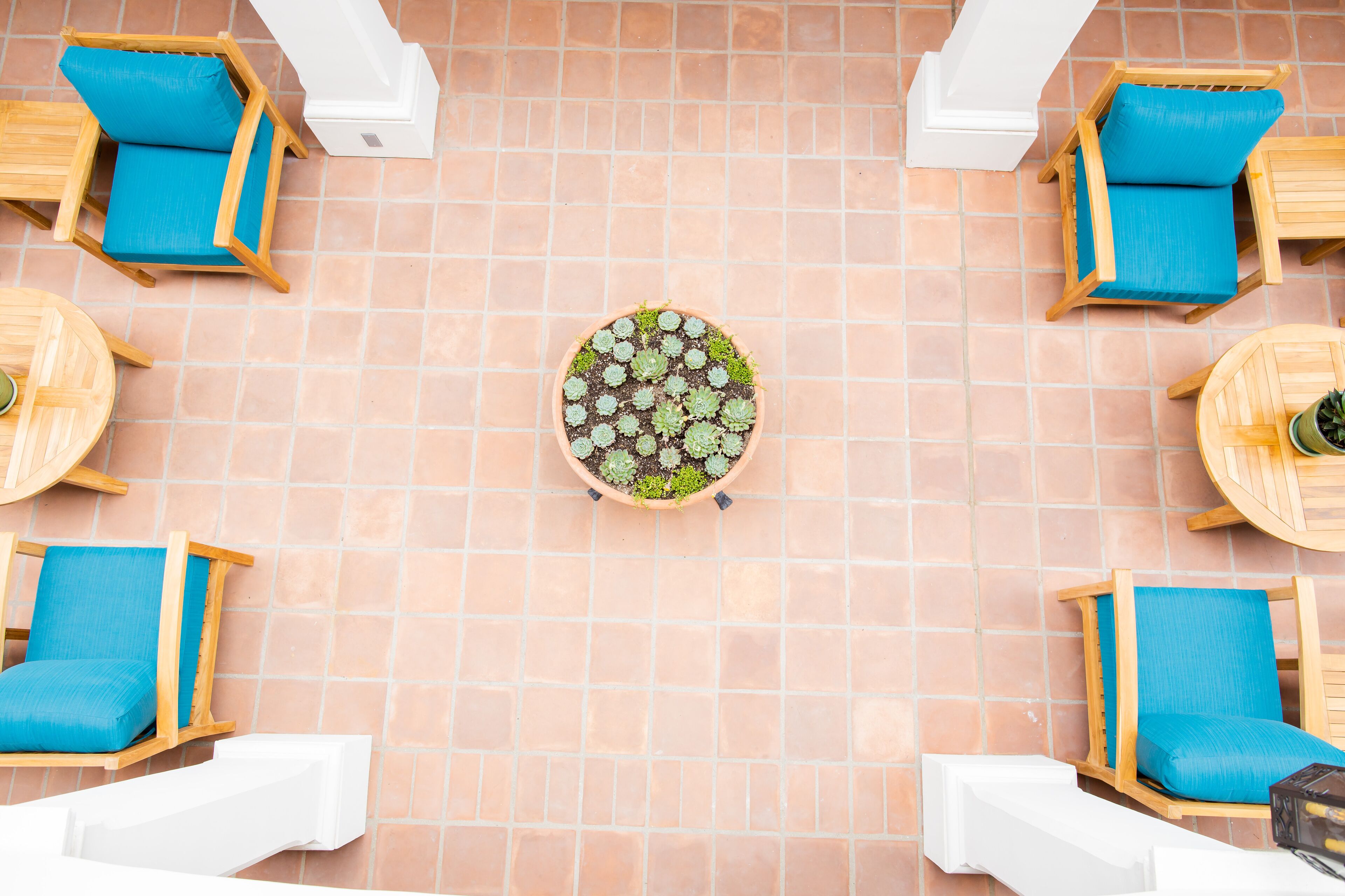 A tiled patio with four wooden chairs and tables, each with blue cushions, arranged around a central potted plant.