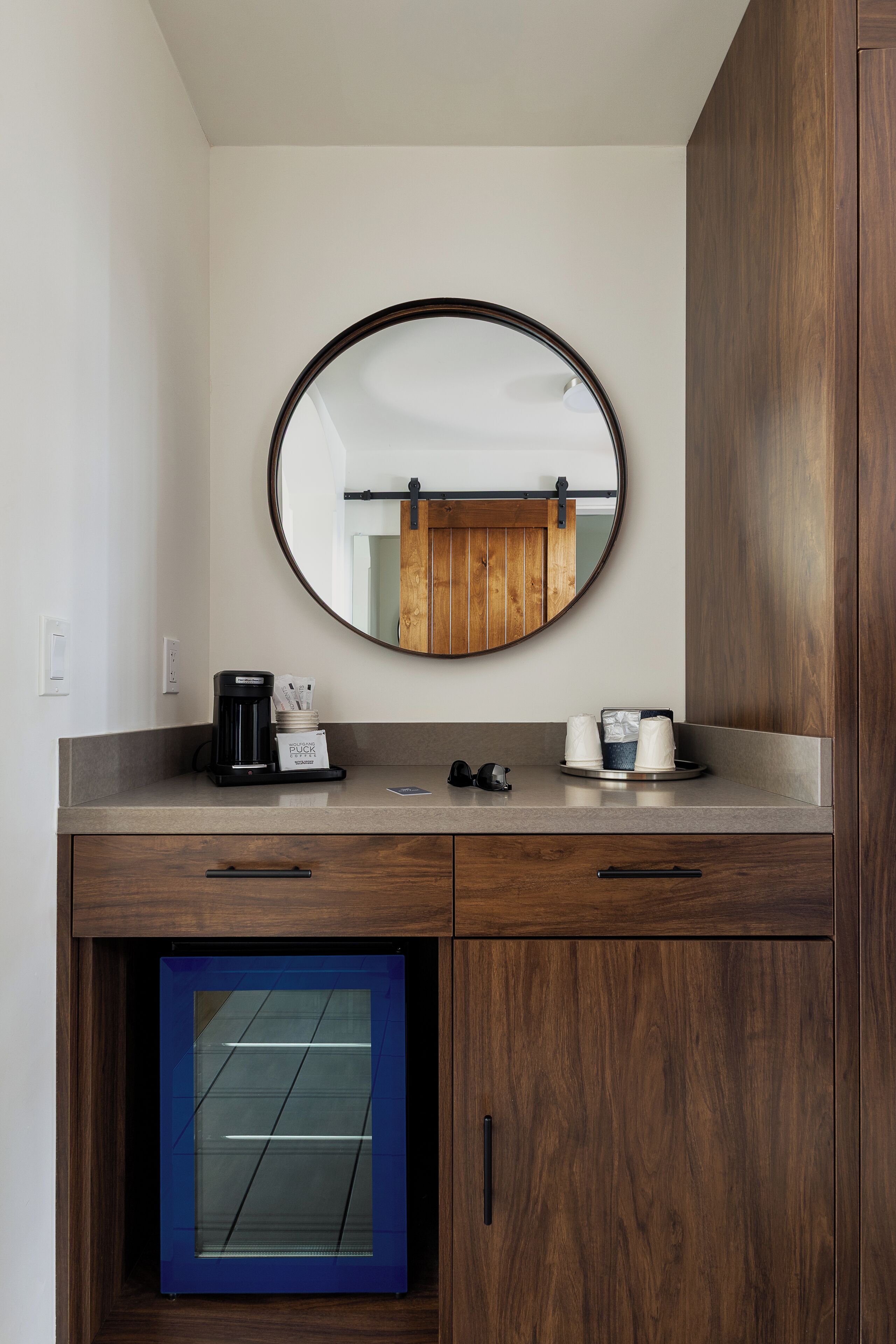 A bathroom with a round mirror, a coffee maker, a cup, and a small cabinet.