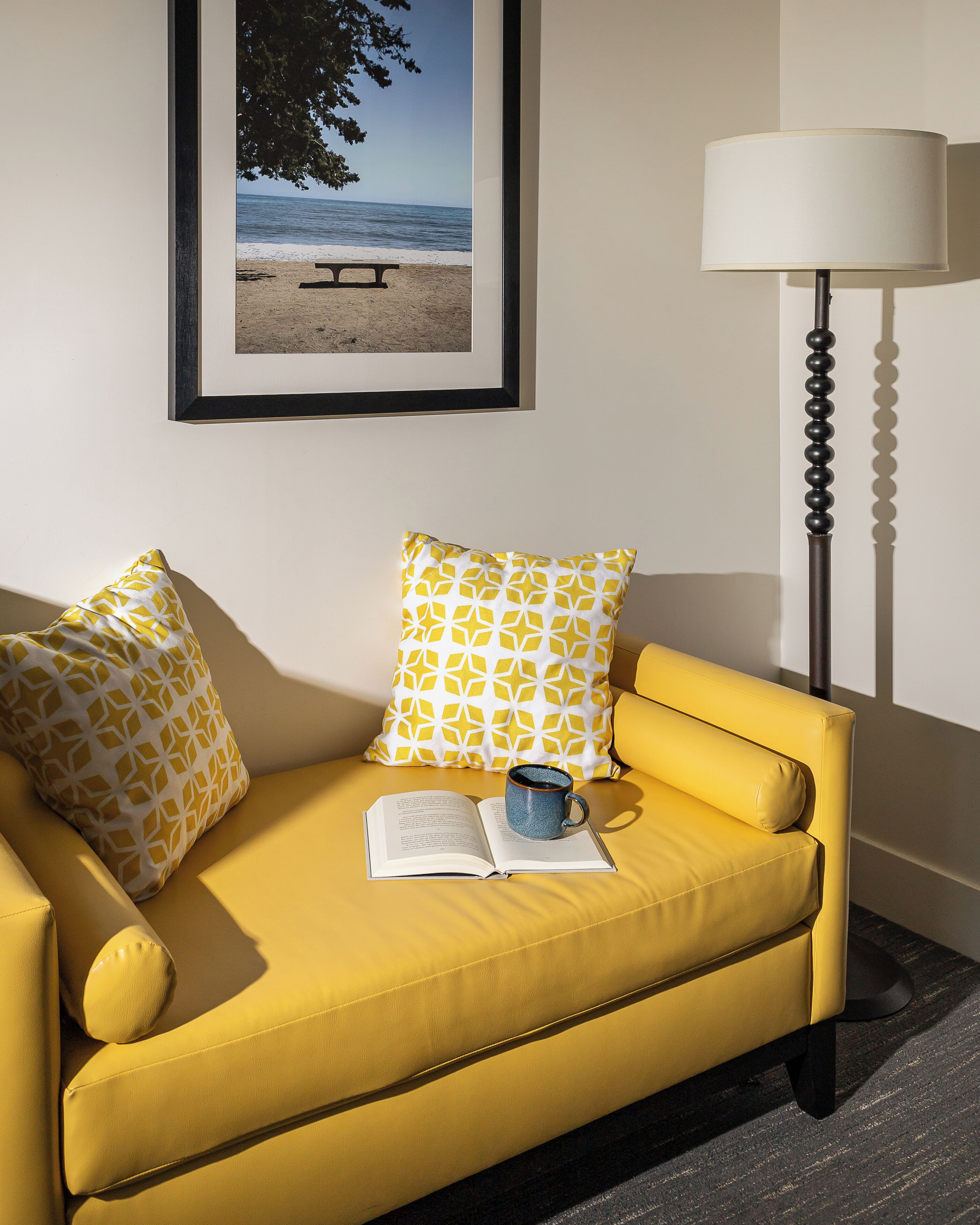 A yellow sofa with patterned pillows, a book, and a mug, a framed picture of a beach, and a floor lamp.