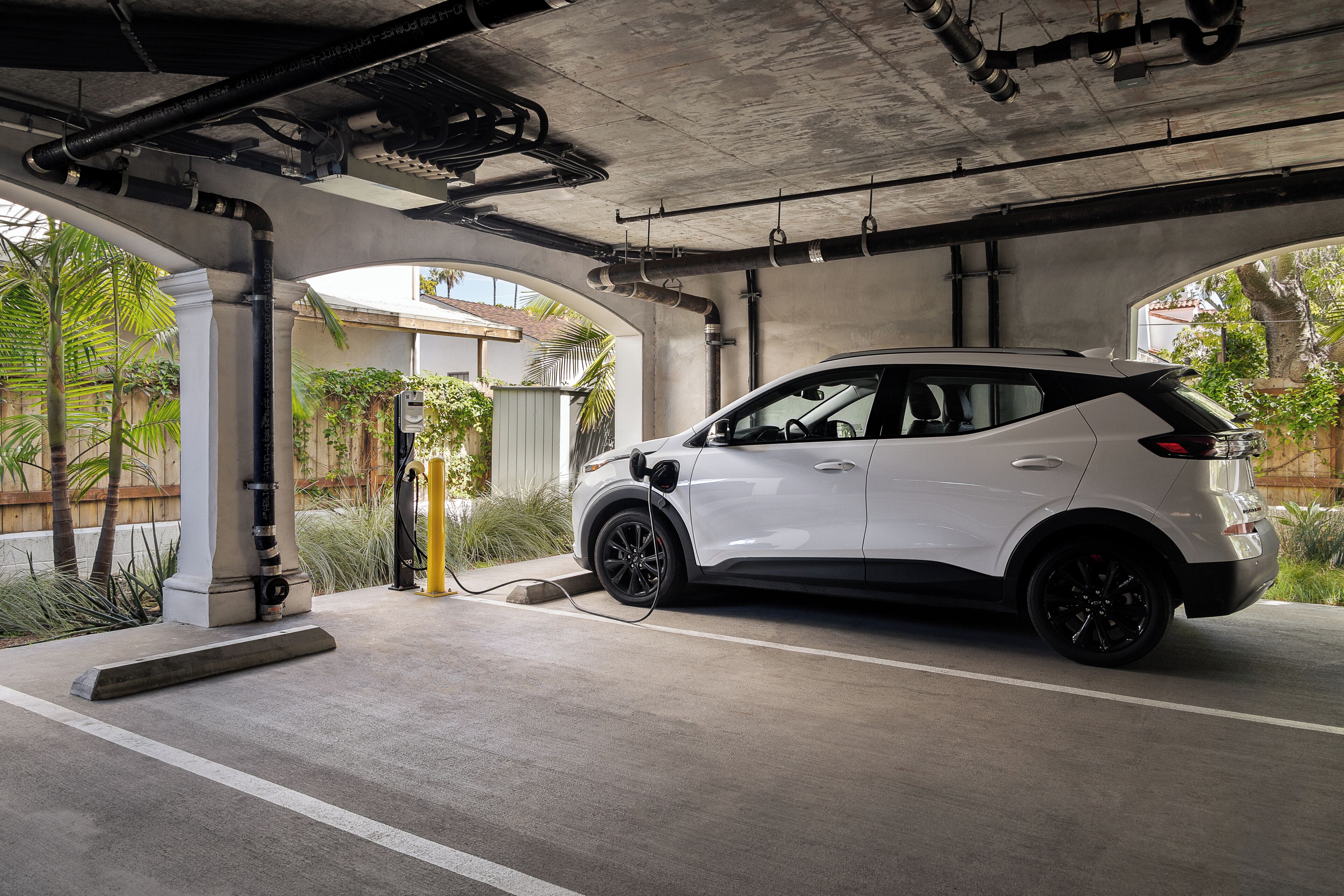 En vit SUV står parkerad i ett överbyggt parkeringsområde med betongpelare och rör som löper längs taket.